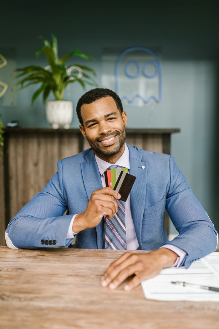 about-01 A confident businessman in a blue suit holds and displays credit cards while smiling at a desk.